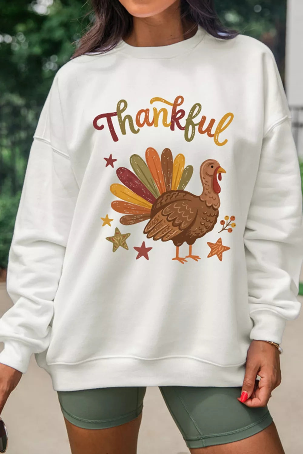 Modell in Olivshorts trägt weißen Crewneck-Pullover mit buntem Truthahn-Print und 'Thankful'-Aufdruck