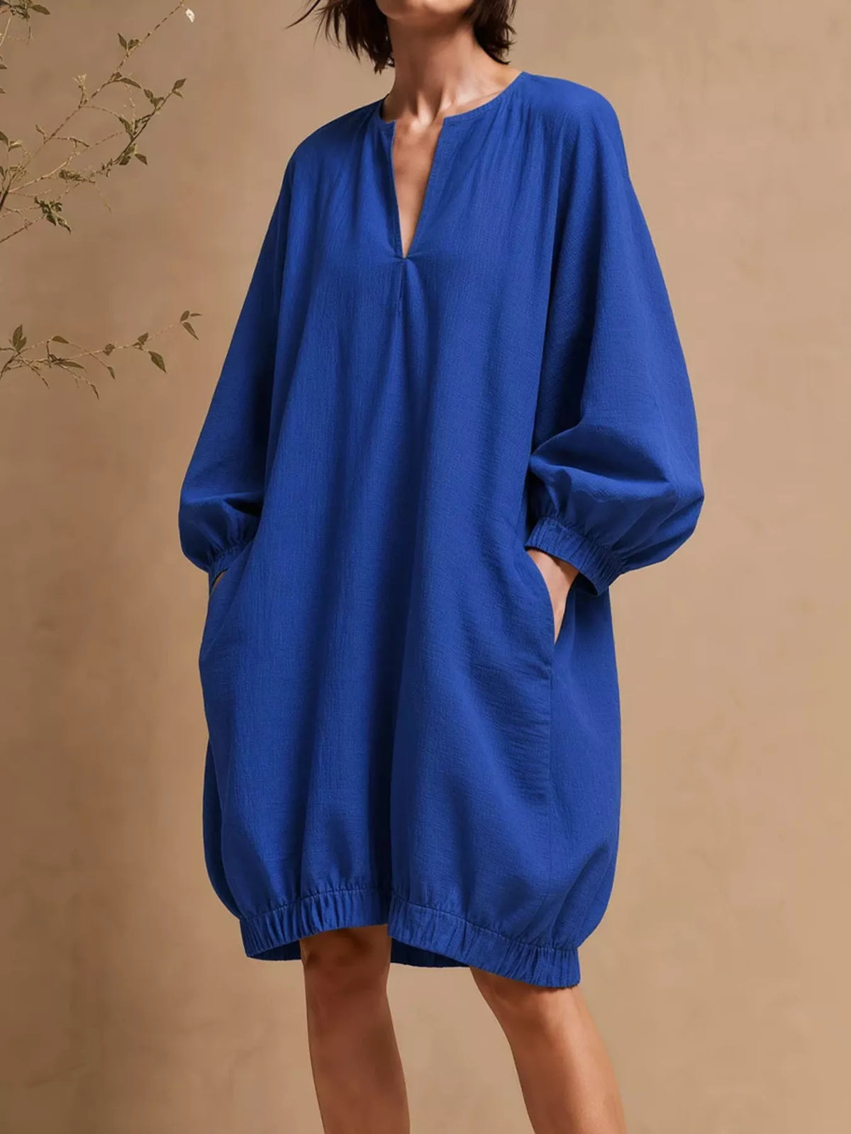 Mujer con vestido mini azul real de manga globo, escote V y bolsillos de pie contra fondo beige cálido
