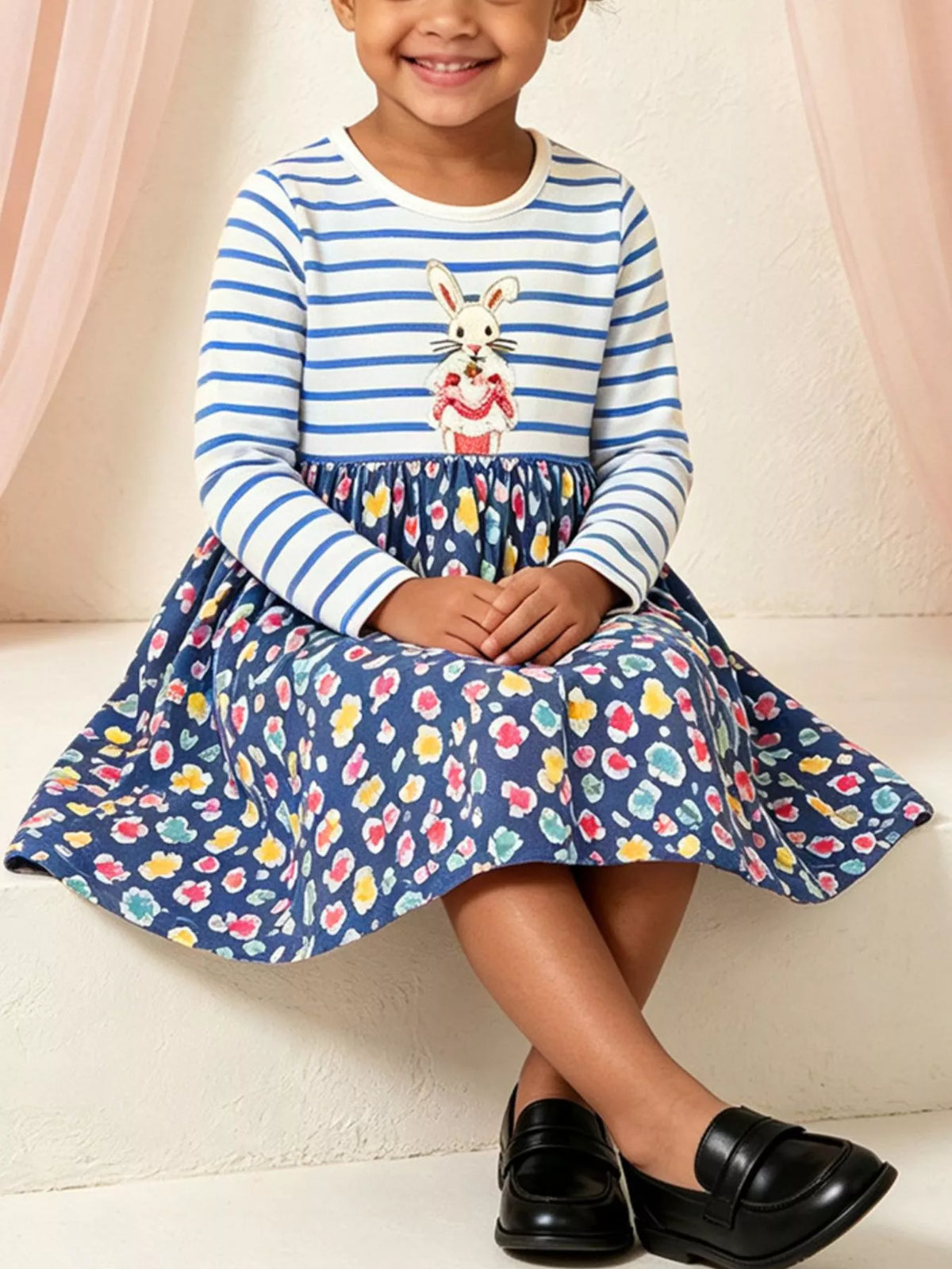 Fille en robe à rayures avec lapin brodé blanc et jupe fleurie marine assise sur des marches