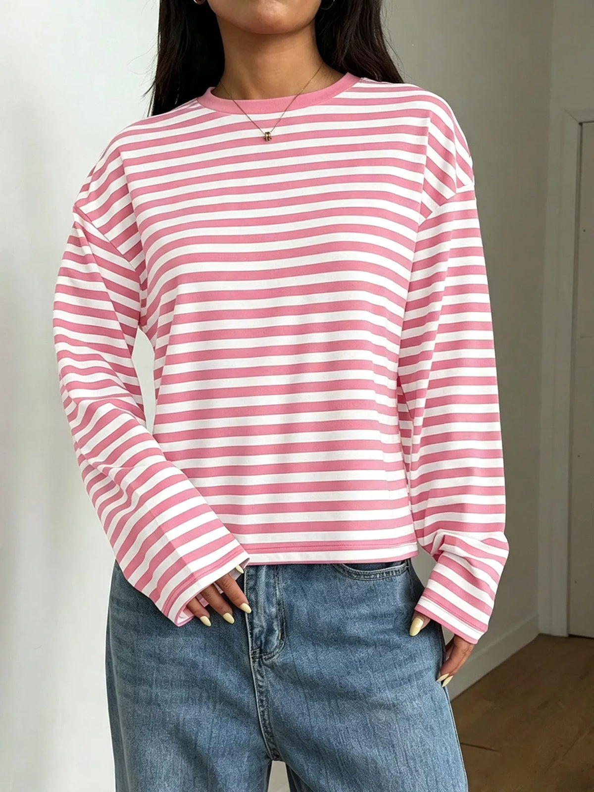 Modella in top maniche lunghe tondo a righe orizzontali rosa e bianche abbinato a jeans lavaggio chiaro su sfondo neutro