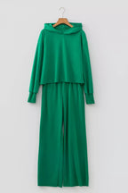Conjunto de dos piezas verde colgado en percha sobre pared blanca: sudadera corta y pantalón amplio para descanso diario.