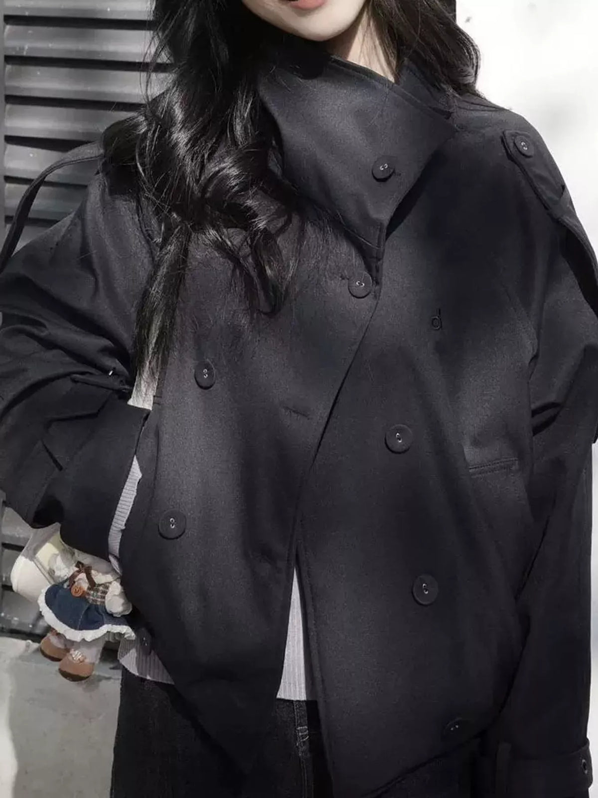 Woman in black double-breasted cropped trench jacket with stand-up collar and shoulder epaulettes against slatted background