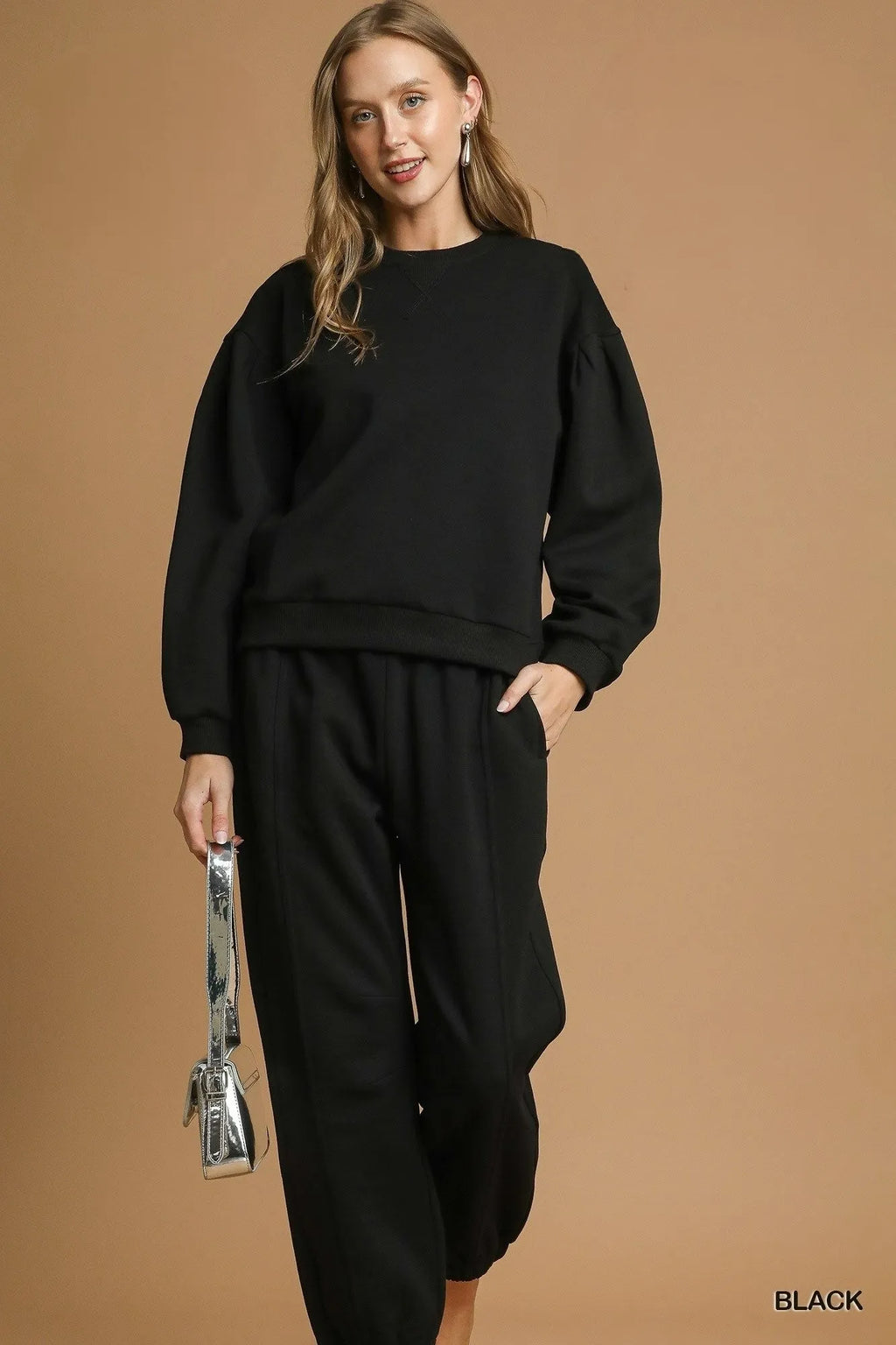 Model in black pleated round neck sweatshirt paired with matching joggers and holding a silver handbag, illustrating a cozy