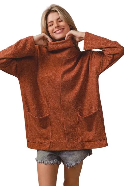 Blonde model in rust orange ribbed turtleneck sweater adjusting collar with hands, wearing denim shorts against beige